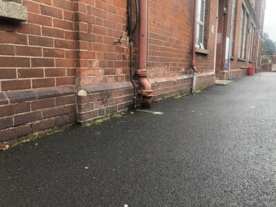 Cast iron rainwater downpipe on a red brick elevation beside a tarmac path