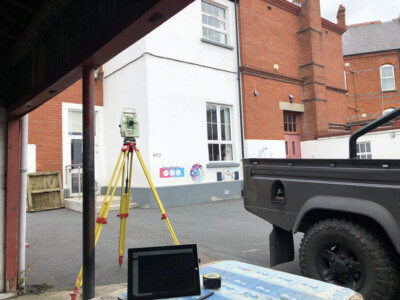 Total station on a tripod in front of a rendered and red brick building