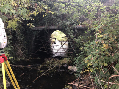 Stone arch bridge over a stream in dense woodland, tripod leg in view