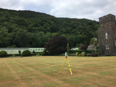 Survey tripod on a lawn below a wooded hillside beside a stone tower